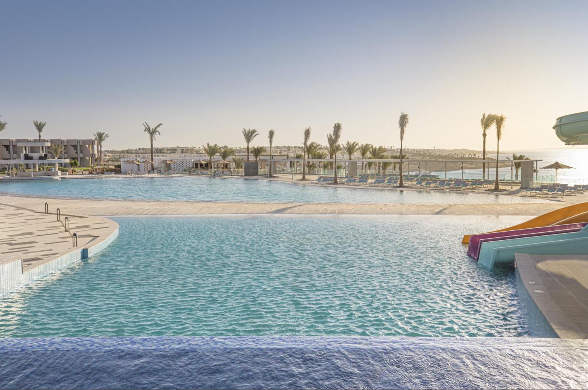 V Hotel Sharm El Shiekh Εξωτερικό φωτογραφία The photo shows a serene outdoor pool area with a scenic view. There is a large pool with crystal-clear water, surrounded by palm trees. On one side, there's a water slide with colorful sections poking out of the pool. In the background, you can see 
